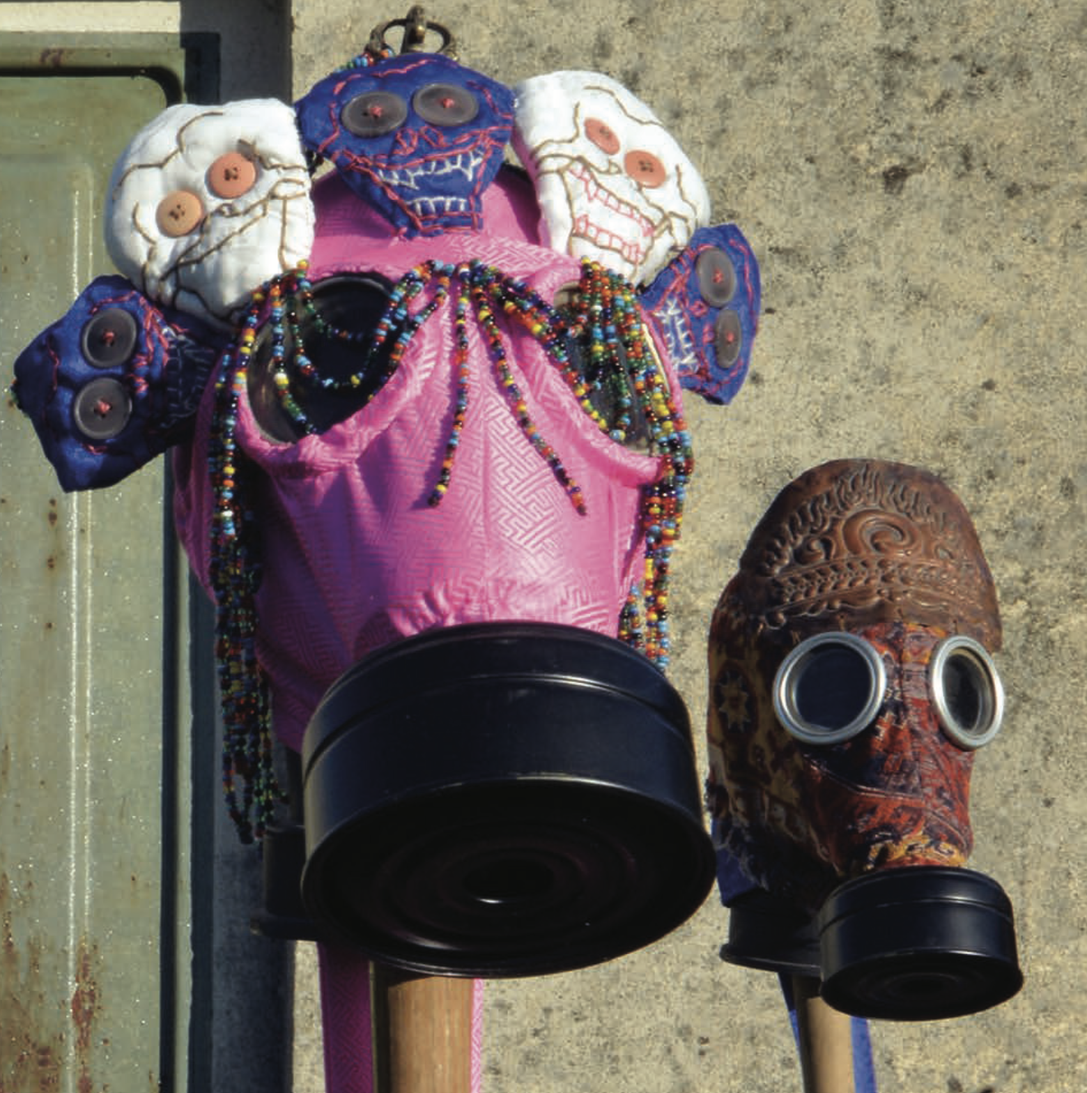 Two gas-mask sculptures side by side: one wrapped in pink fabric with beaded strands and skull-like forms, the other covered in patterned fabric with round glass lenses