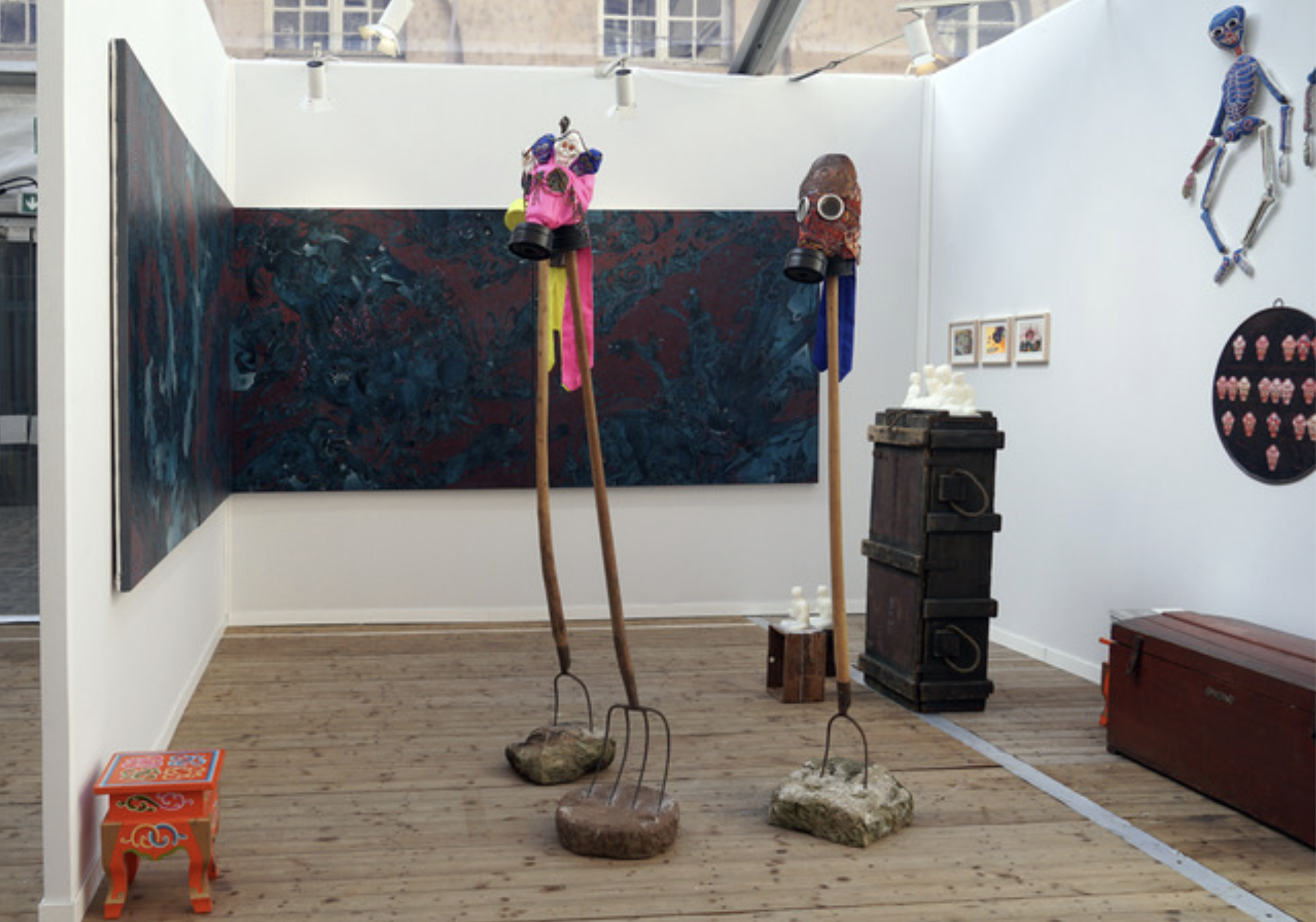 THEgallery booth with a large blue-and-red painting, two tall gas-mask sculptures on poles, a dark crate topped with small white figures, and works on the white walls